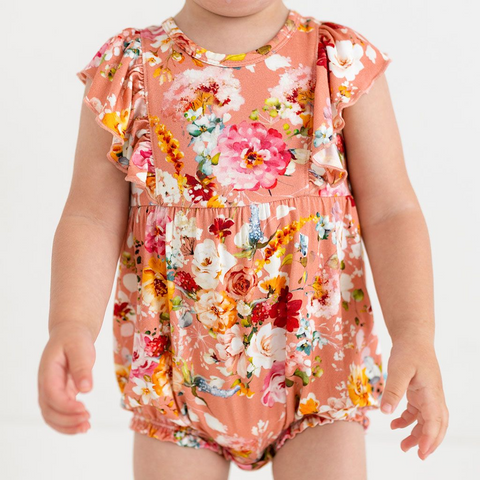 toddler wearing a flutter sleeve romper. Print is orange, red, yellow and white floral on a peachy background.