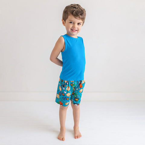 Boy standing in tank top and shorts. Tank top is solid blue and shorts are a pattern of cartoonish red and green tractors, bails of hay and farm tools are on a blue background.