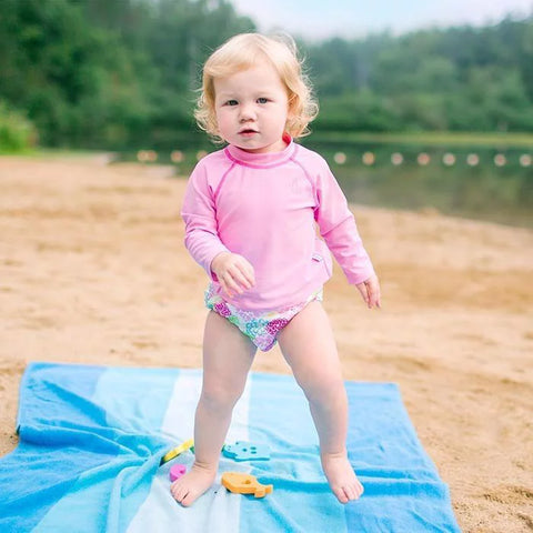 Toddler standing on a blue beach towel wearing a pink long sleeve rash guard and a floral swim bottom.