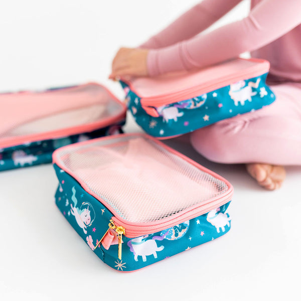 Girl unzipping travel cubes. Pack of 3 zippered travel cubes stacked.  Print is unicorns in space. Blue background with peachy pink trim.