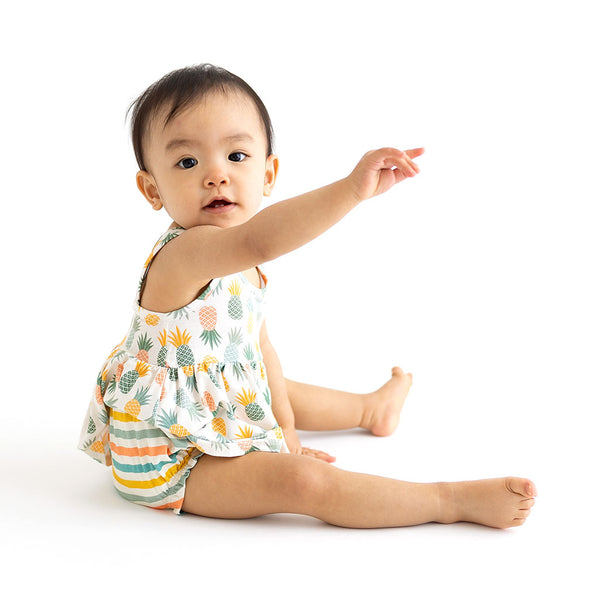 Child sitting in Peplum top is sleeveless and has Pineapple print. Light Green, Dark green, Orange, and Yellow Pineapples are repeated all over gown. It comes with a pair of Bummies bottoms that have coordinating colors in stripes.