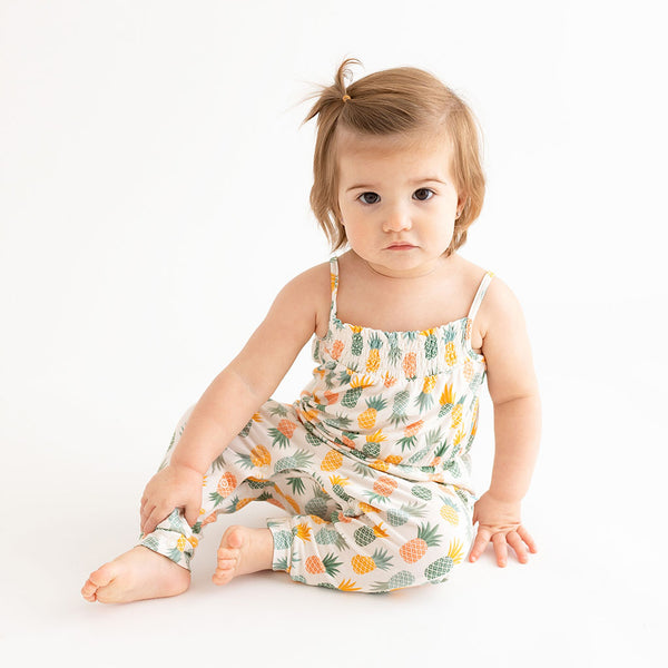 Toddler sitting. She is wearing Jumpsuit has a white background with Light Green, Dark Green, Yellow, and Orange Pineapples all over it.