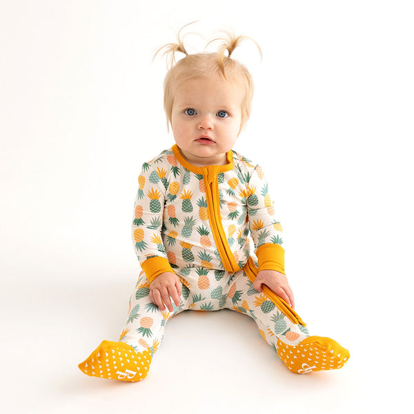 Toddler sitting down in the One Piece. The Bottom of the Footies are showing. They are Bright orange with little foam white hearts to prevent slipping when walking. This is the Pineapple print.  Light Green, Dark green, Orange, and Yellow Pineapples are repeated all over gown.