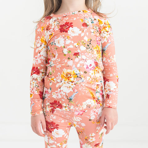 Girl wearing long sleeved two piece pajama. Print is red, white and yellow flowers on a peach background.