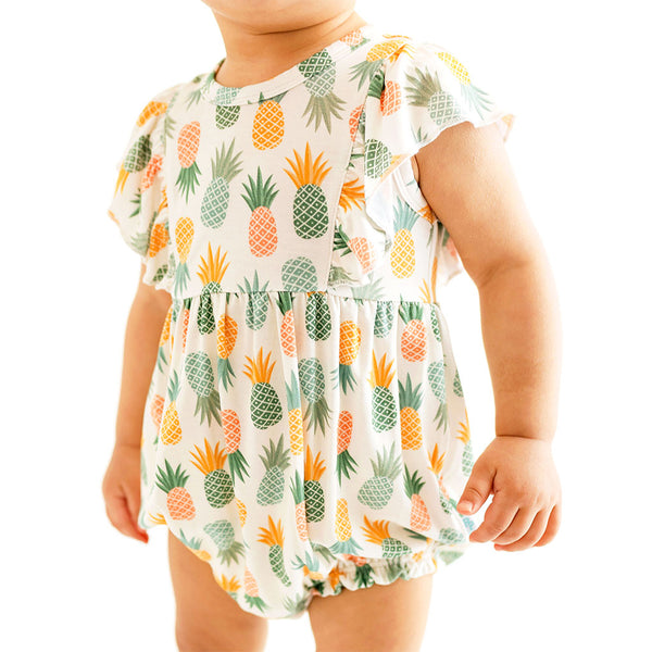 Close up of child standing in the Flutter Sleeve Romper with easy diaper change snaps. This is the This print has a white background with Pineapple print. Light Green, Dark green, Orange, and Yellow Pineapples are repeated all over with a white background
