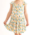 Close up of girl wearing Flutter Sleeve Tiered dress has a white background with Pineapple print. Light Green, Dark green, Orange, and Yellow Pineapples are repeated all over with a white background