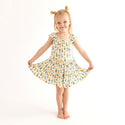 Girl standing and holding the edges of her dress. She is wearing Flutter Sleeve Tiered dress has a white background with Pineapple print. Light Green, Dark green, Orange, and Yellow Pineapples are repeated all over with a white background