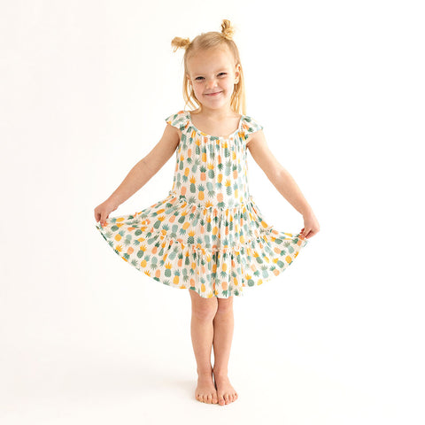 Girl standing and holding the edges of her dress. She is wearing Flutter Sleeve Tiered dress has a white background with Pineapple print. Light Green, Dark green, Orange, and Yellow Pineapples are repeated all over with a white background