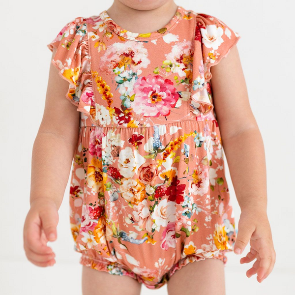 toddler wearing a flutter sleeve romper. Print is orange, red, yellow and white floral on a peachy background.