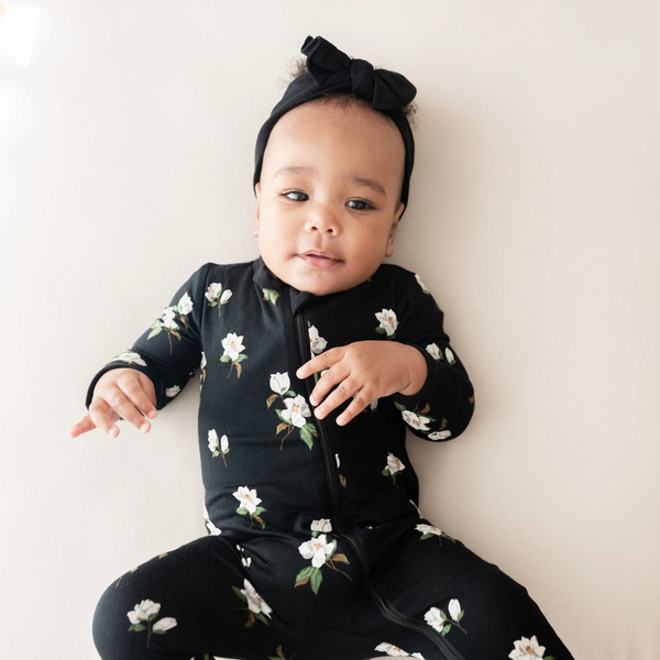 baby wearing a footie pj in An all-over floral print featuring small white magnolias with their leaves and stems contrasting vividly against a black background color called Midnight.