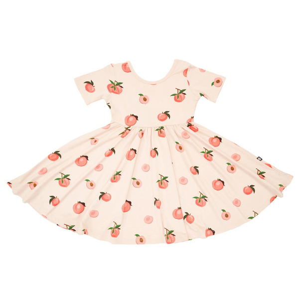 An all-over print featuring shiny whole peaches on the branch, whole peaches with their stems and leaves, halved peaches with their leaves and pits, and halved peaches without their pits, over a pinkish beige background color called Porcelain.