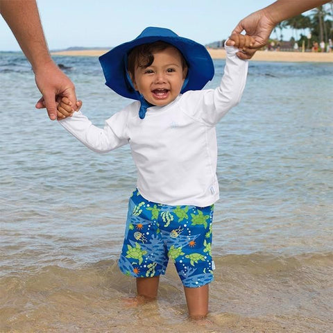 Toddler wearing a white long sleeve rash guard and a blue sun hat while playing at the beach.