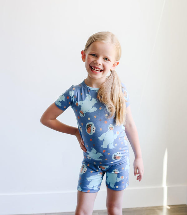 Blonde girl with a pony tail standing with hand on hip wearing two piece short pajama set. Blue background with cookie monster and cooke monster heads.
