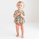 Little Girl standing in a Two-Piece outfit.  Includes a Peplum Top. The Print is a floral on a cream background. The Flowers are single daisies on stems and vary in color. Light Blue, Yellow, light orange, and pink. The Bloomer bottoms are a red and white gingham print and have adorable ruffles on the back.