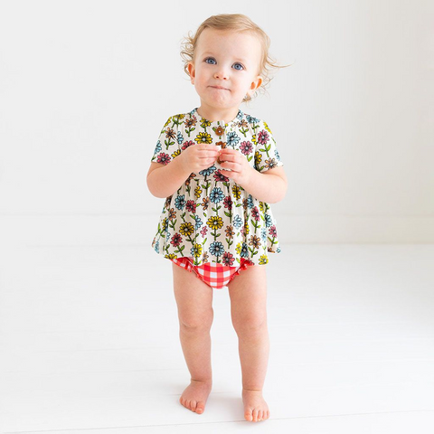 Little Girl standing in a Two-Piece outfit.  Includes a Peplum Top. The Print is a floral on a cream background. The Flowers are single daisies on stems and vary in color. Light Blue, Yellow, light orange, and pink. The Bloomer bottoms are a red and white gingham print and have adorable ruffles on the back.