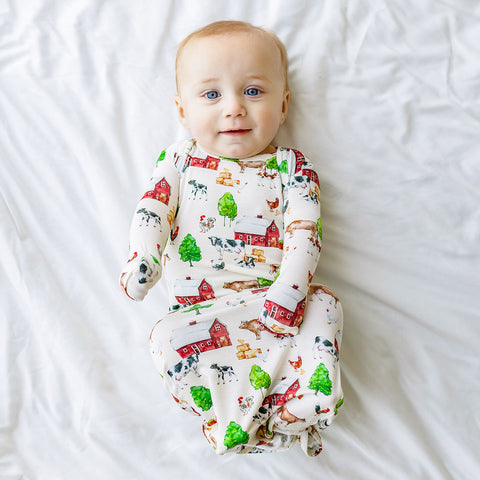 Baby in knotted gown with mitts over hands. Country farm print on a cream background. 