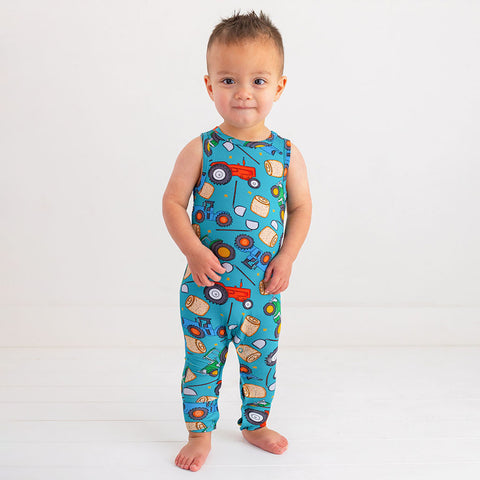Toddler boy in a racerback long pant romper. Pattern of cartoonish red and green tractors, bails of hay and farm tools are on a blue background.