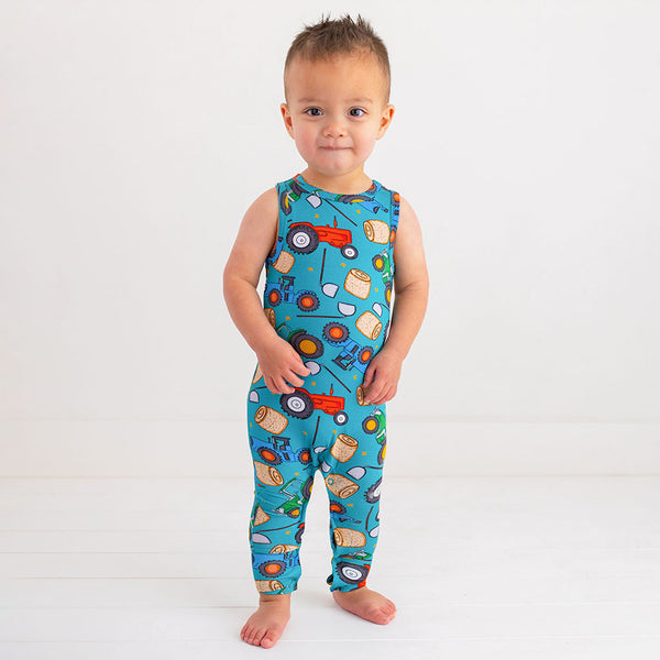 Toddler boy in a racerback long pant romper. Pattern of cartoonish red and green tractors, bails of hay and farm tools are on a blue background.