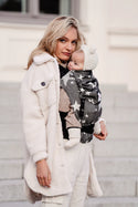 Woman in white coat wearing a babycarrier. Carrier is black with white large stars.