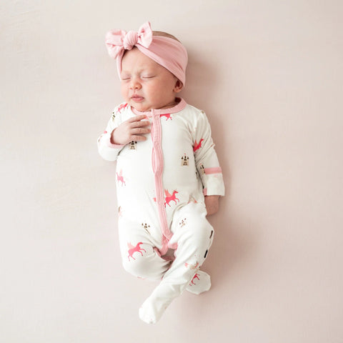 Infant wearing white romper with pink unicorns and castles on it. Arm cuffs, neck and zipper trim are pink.