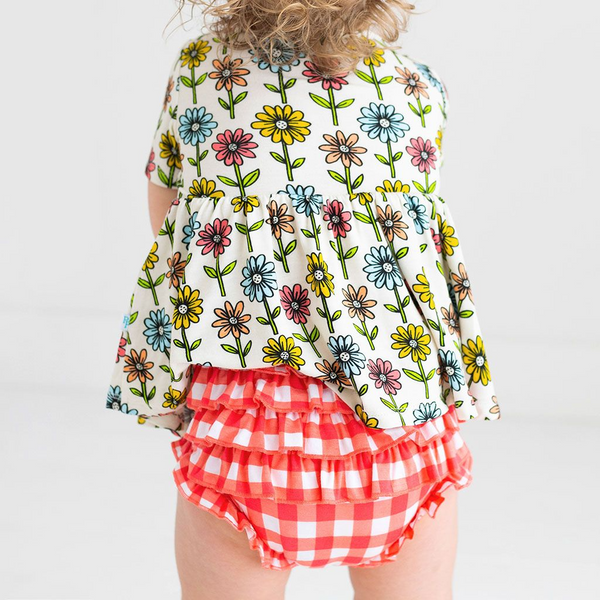 Little Girl standing with her back facing the camera in a Two-Piece outfit.  Includes a Peplum Top. The Print is a floral on a cream background. The Flowers are single daisies on stems and vary in color. Light Blue, Yellow, light orange, and pink. The Bloomer bottoms are a red and white gingham print and have adorable ruffles on the back.