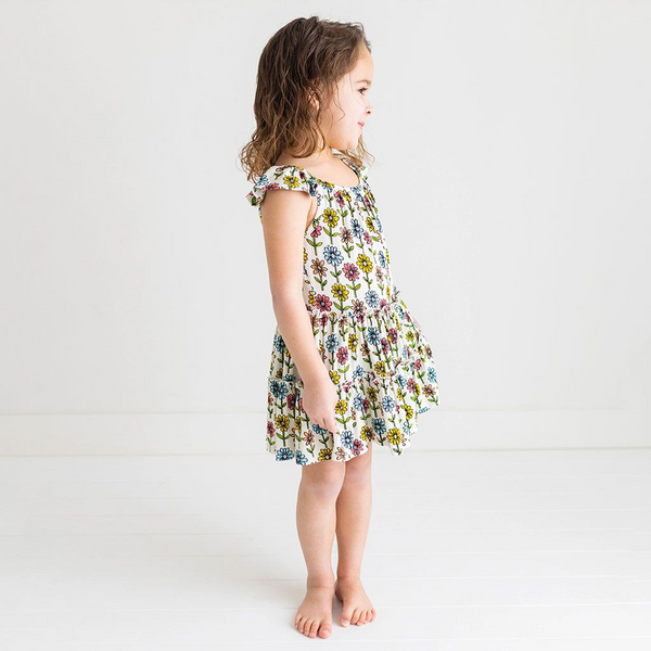 A little girl standing in a 3 Tiered Dress with Flutter Sleeves. The dress Print is a floral on a cream background. The Flowers are single daisies on stems and vary in color. Light Blue, Yellow, light orange, and pink.