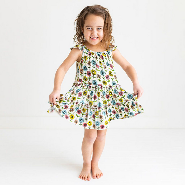 A little girl standing and holding the sides of her dress to show a 3 Tiered Dress with Flutter Sleeves. The dress Print is a floral on a cream background. The Flowers are single daisies on stems and vary in color. Light Blue, Yellow, light orange, and pink.